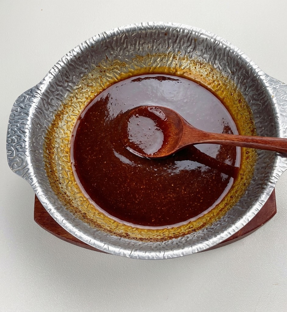 Gotowy ciemny sos miso delikatnie gotujący się w metalowym garnku, wyglądający na gęsty i błyszczący, z drewnianą łyżką spoczywającą na środku.