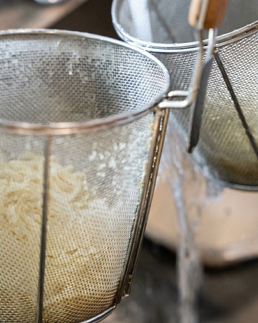 Gruby makaron ramen wewnątrz cylindrycznego sita płukany pod strumieniem bieżącej zimnej wody.