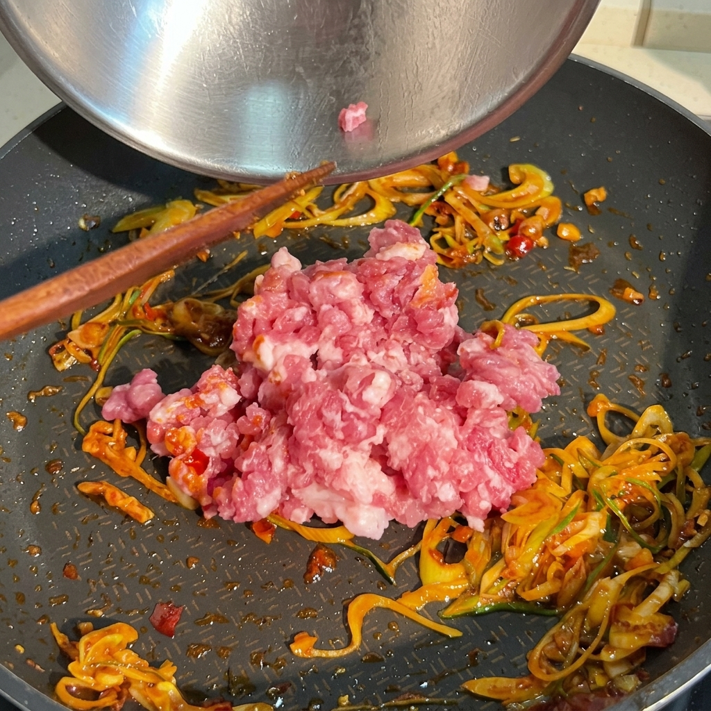 Porcja surowego, różowego mięsa mielonego dodawana na patelnię z podsmażoną zieloną cebulką i czerwonym sosem chili.