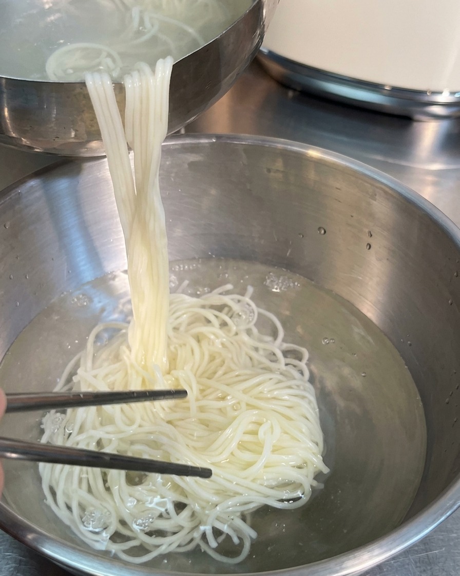 Srebrne pałeczki wyjmujące dużą porcję ugotowanego białego makaronu z metalowej miski z wodą do drugiej miski.