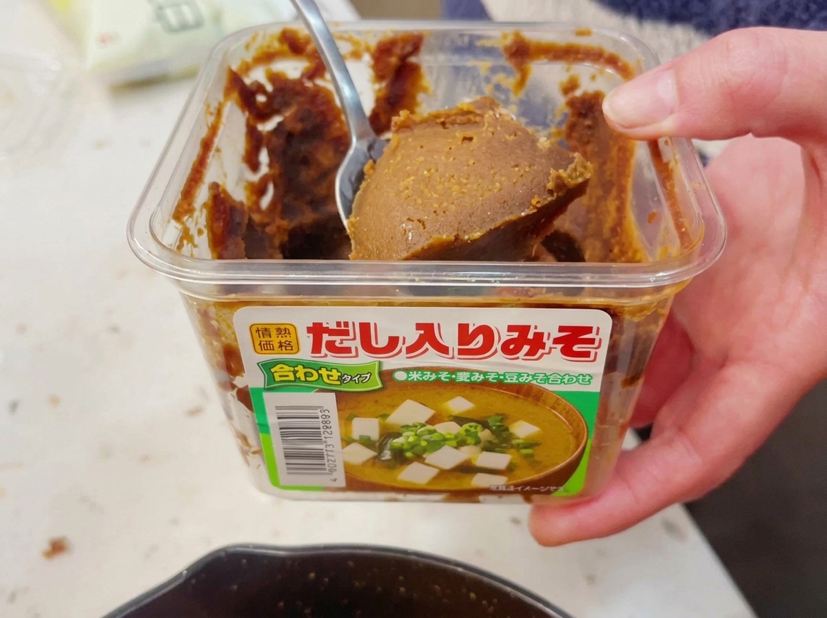 Dłoń nabierająca metalową łyżką obfitą porcję gęstej, brązowej pasty miso z przezroczystego plastikowego pojemnika.