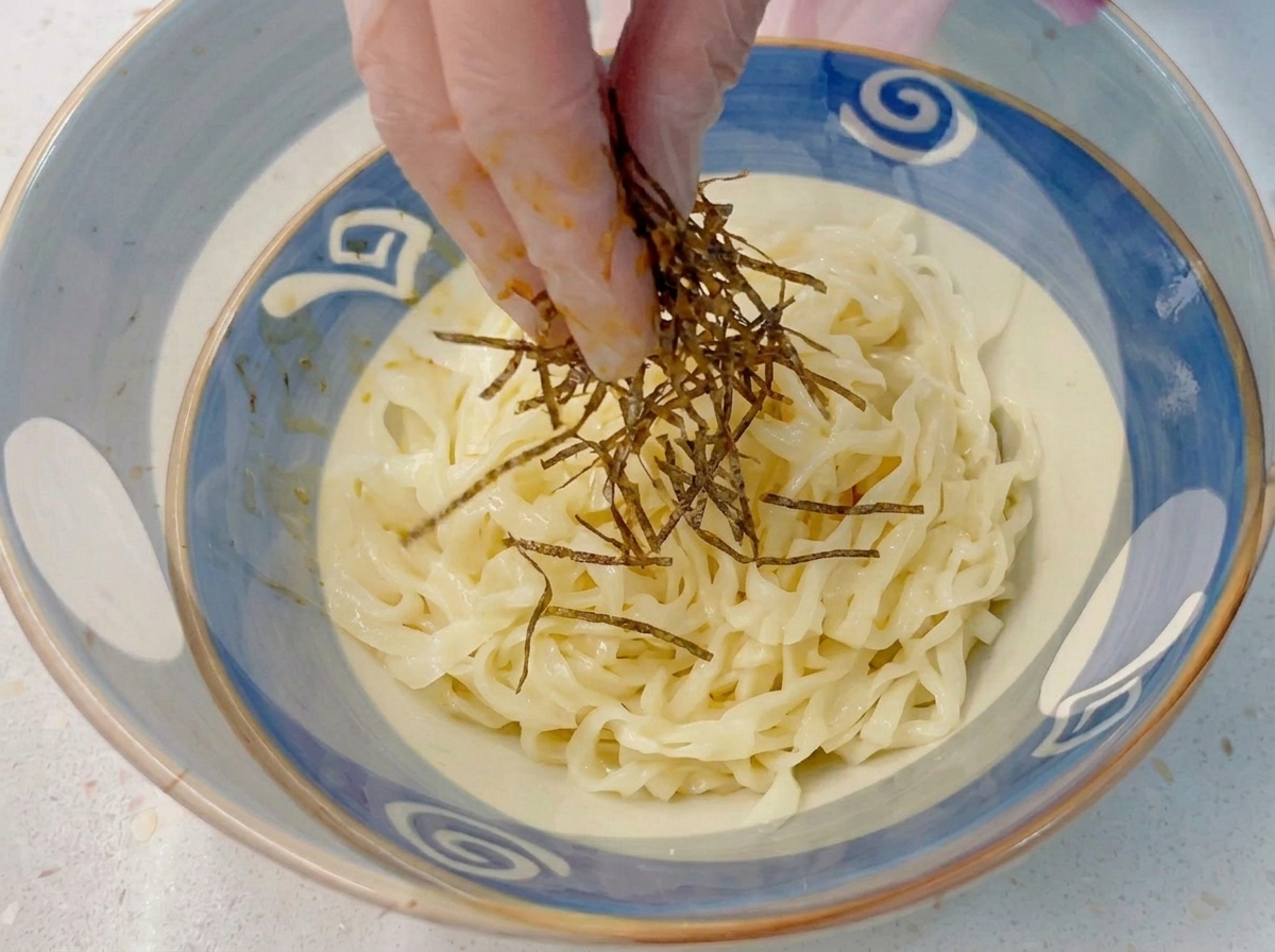 Palce w rękawiczce układające paski wodorostów nori na misce z zimnym, grubym makaronem ramen.