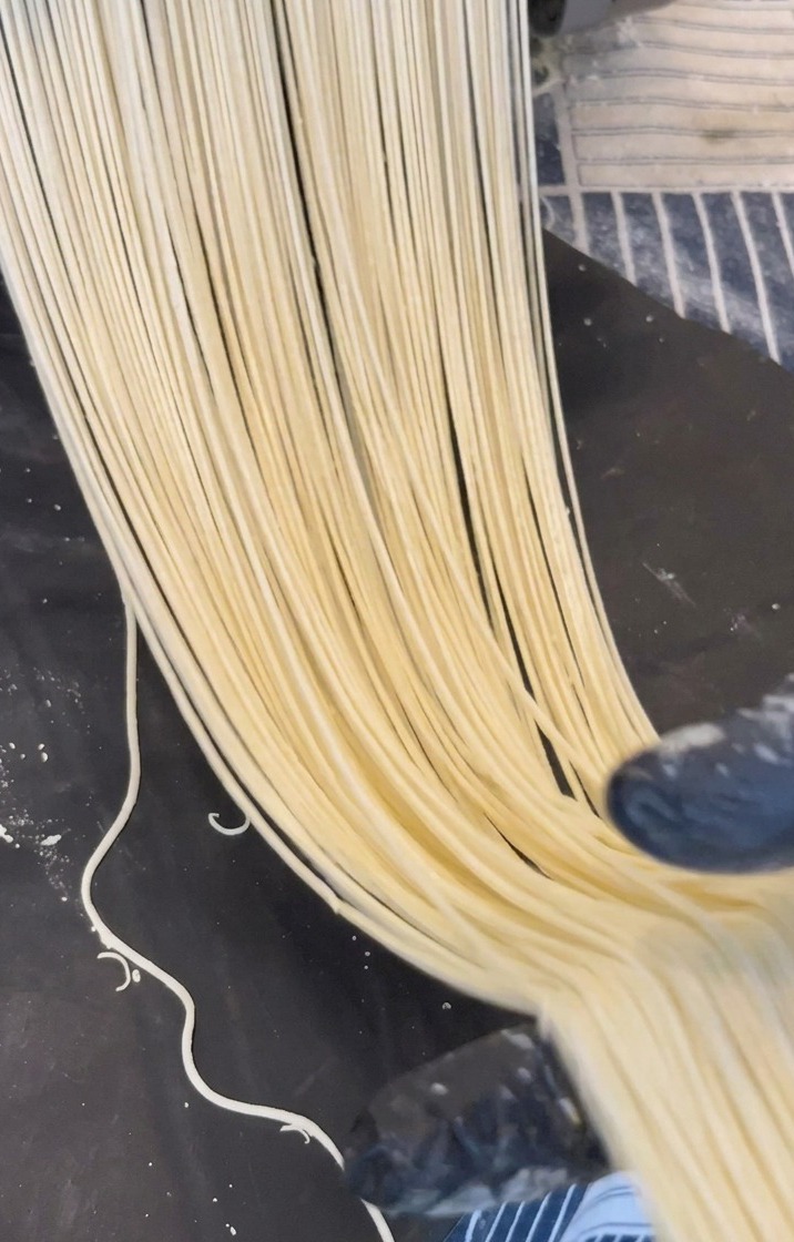 Długie nitki jasnożółtego makaronu ramen wychodzące z metalowej maszynki na ciemny blat.
