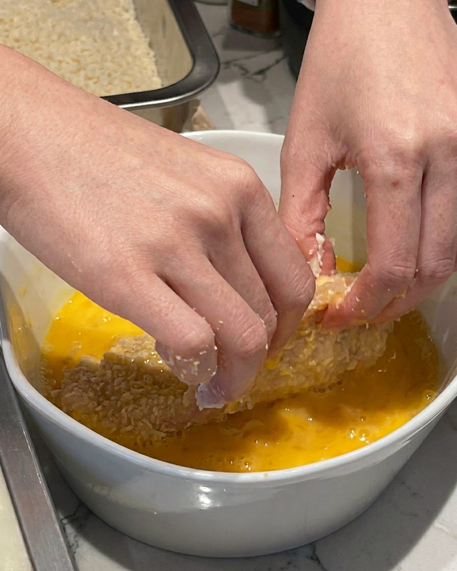 Ręka zanurzająca obtoczony w mące kotlet schabowy w białej ceramicznej misce z roztrzepanym żółtym jajkiem.