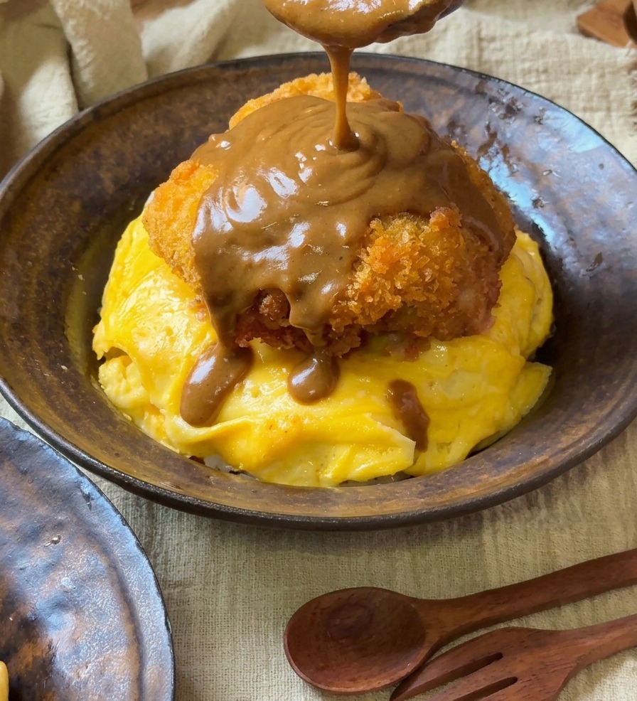 Gęsty, gładki brązowy sos curry wylewany z drewnianej łyżki na złocisty, panierowany kotlet wieprzowy spoczywający na omlecie tornado.