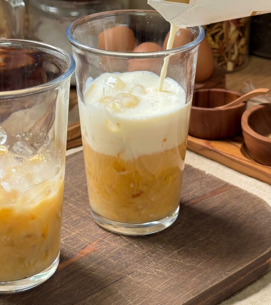 Zimne mleko nalewane z kartonu do wysokiej szklanki wypełnionej lodem i żółtym puree bananowym.