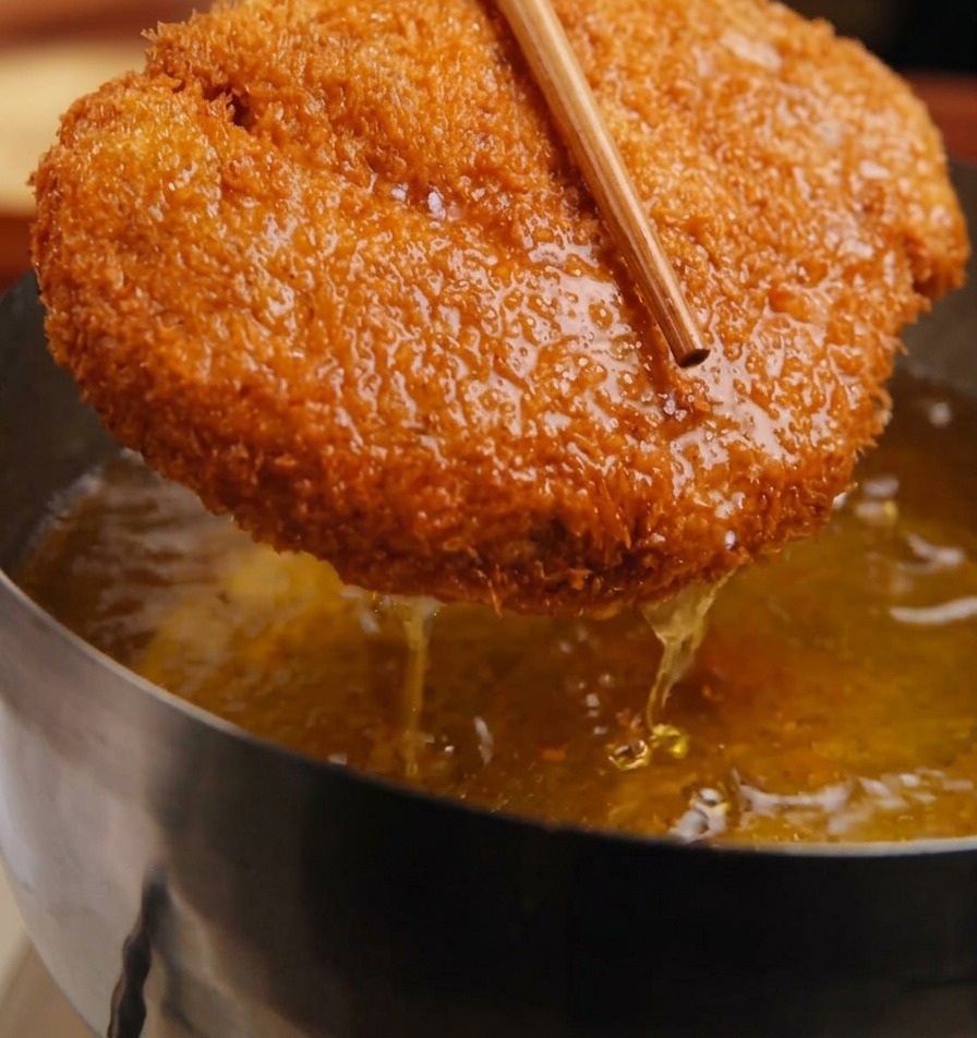 Złocisty kotlet wieprzowy w panierce panko trzymany za pomocą drewnianych pałeczek nad garnkiem z bulgoczącym olejem.