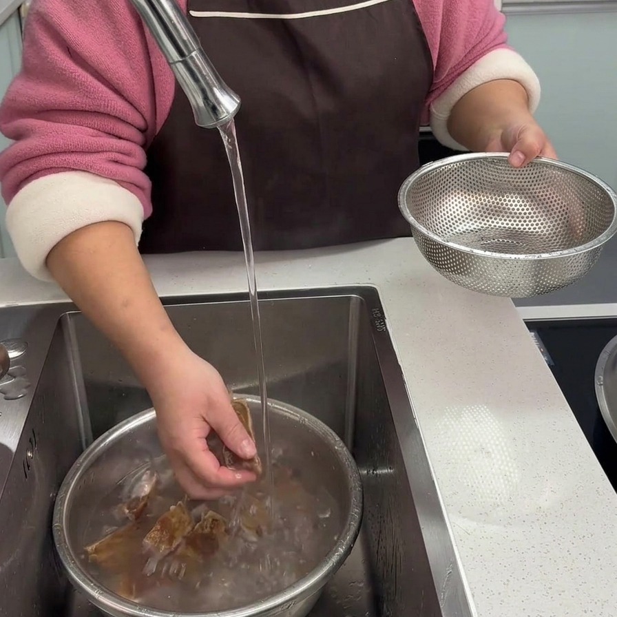 Dłonie dokładnie myjące obgotowane kawałki żeberek pod strumieniem bieżącej czystej wody w zlewie kuchennym.