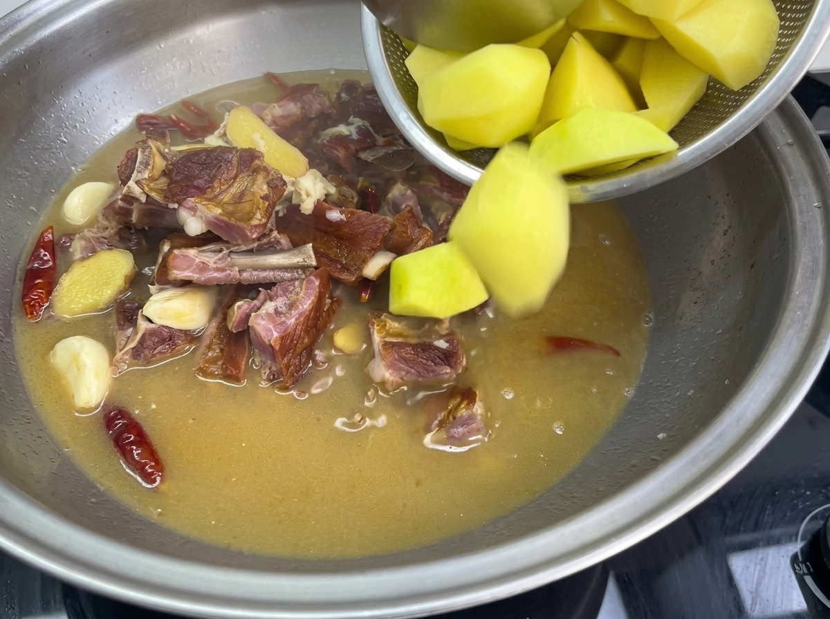 Wrzucanie kawałków złocistych ziemniaków z sitka do dużego metalowego naczynia z gotującym się wywarem, żeberkami, czosnkiem i czerwonym chili.