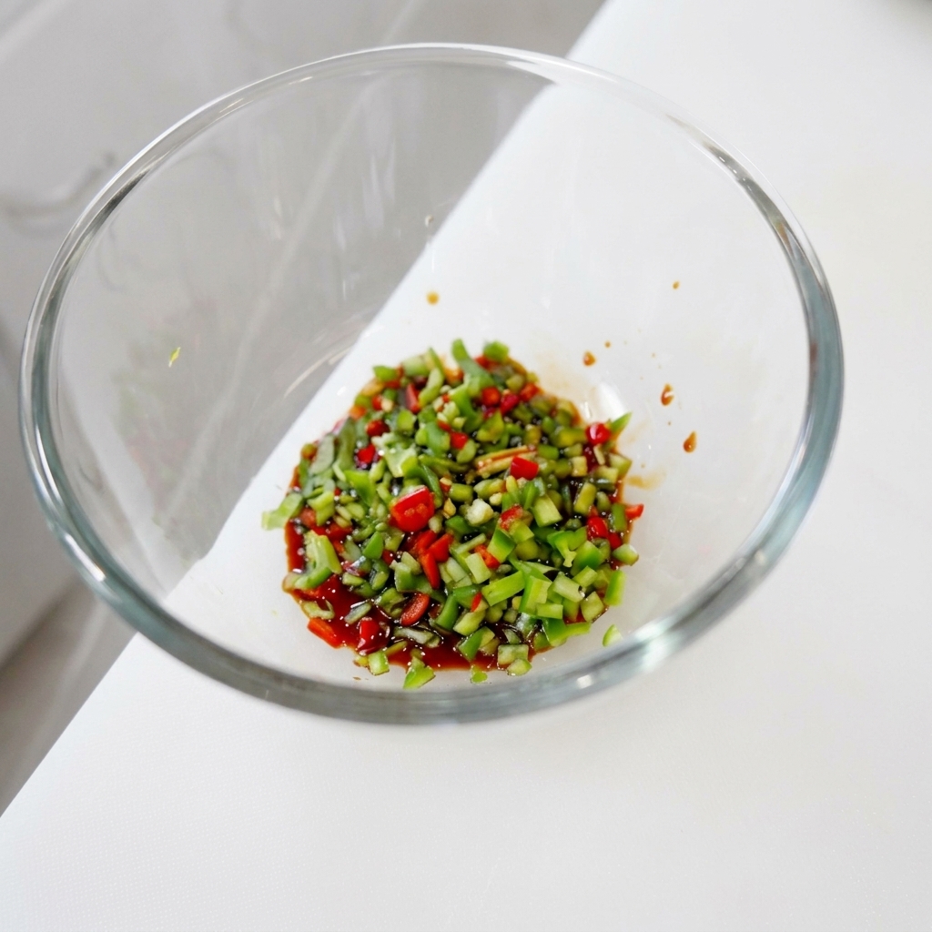Kolorowa mieszanka drobno posiekanych zielonych i czerwonych papryczek chili oraz kolendry zanurzona w ciemnej, błyszczącej płynnej bazie przyprawowej w szklanej misce.