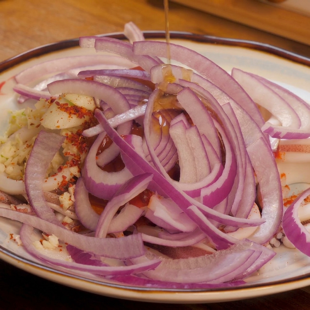 Strumień jasnego oleju sezamowego i ziaren sezamu lądujący na kolorowym stosie cebuli, chili i kalmarów.