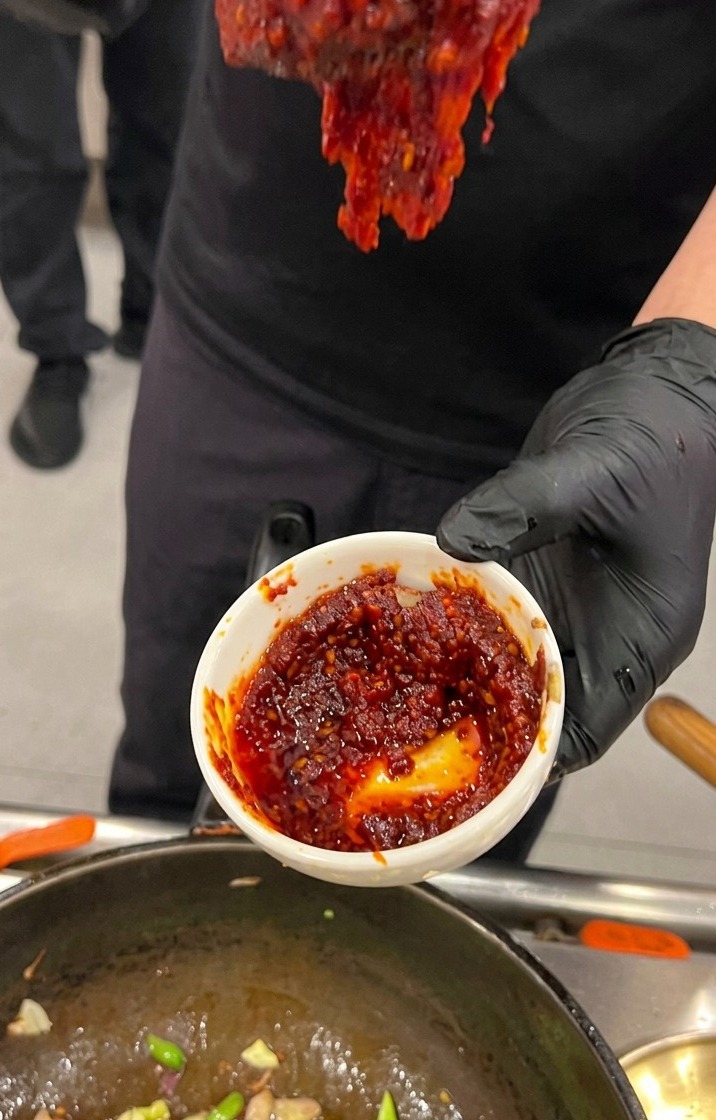 Dłoń w czarnej rękawiczce trzymająca małą białą miseczkę z gęstym, intensywnie czerwonym koreańskim sosem chili nad wokiem.