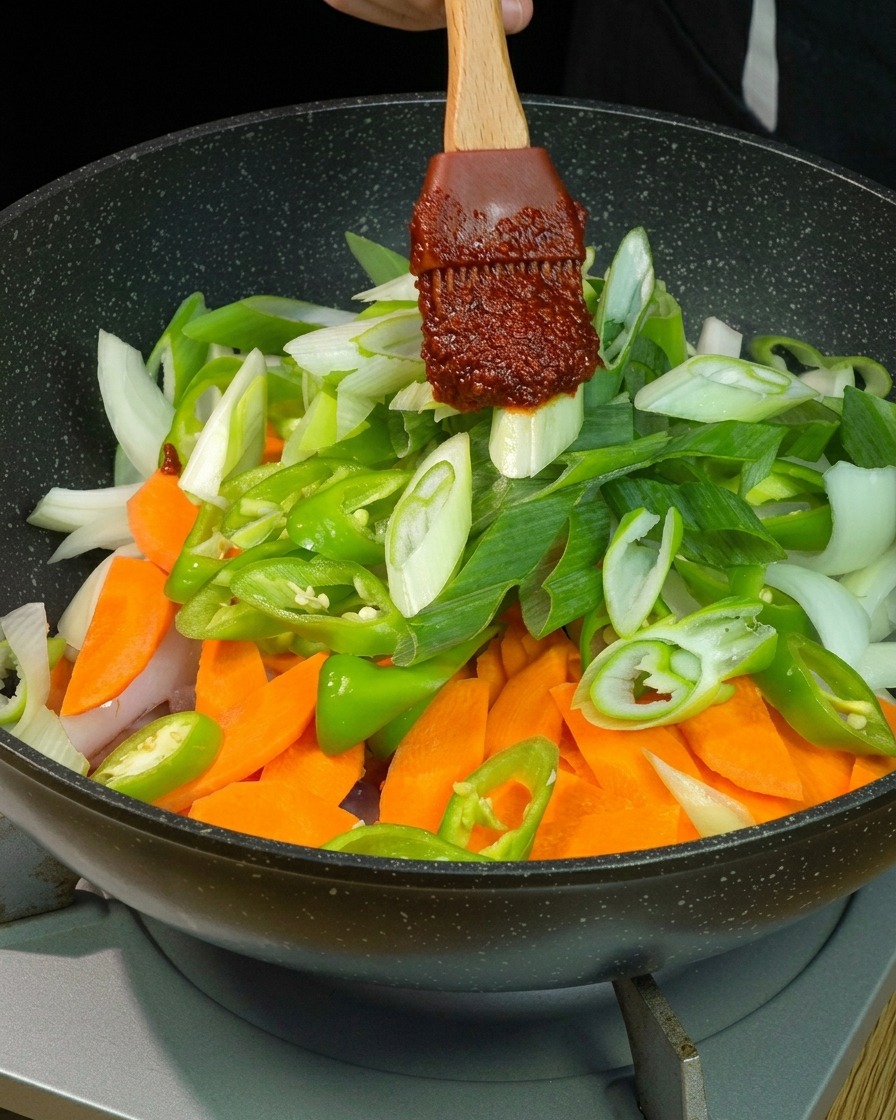 Porcja gęstego, ciemnoczerwonego sosu chili na drewnianej łopatce nakładana na kolorowe warzywa na patelni.