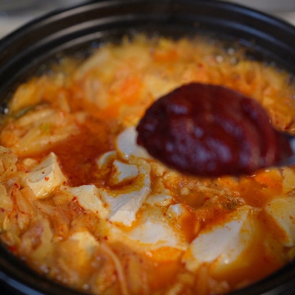 Łyżka z porcją gęstej, ciemnoczerwonej koreańskiej pasty chili nad gotującą się zupą z kimchi i tofu.