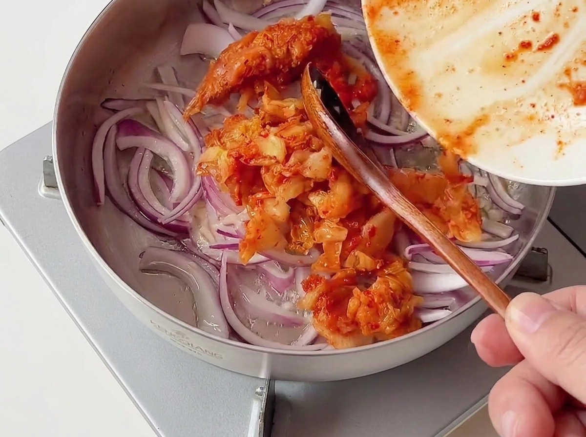 Porcja posiekanego, czerwonego kimchi zsuwana z talerza na patelnię z zeszkloną cebulą i czosnkiem.