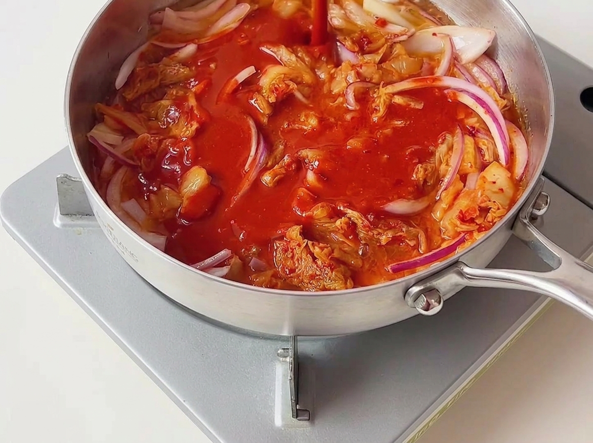 Jaskrawoczerwony sos chili wylewany na podsmażone kimchi i czerwoną cebulę na srebrnej patelni.