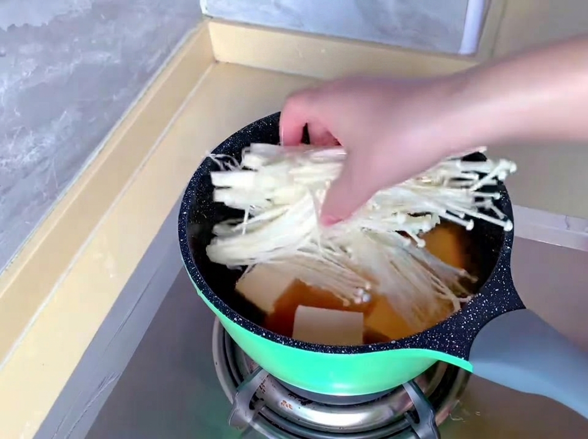 Dłoń wkładająca pęczek długich, białych grzybów enoki do garnka z bulionem i tofu.