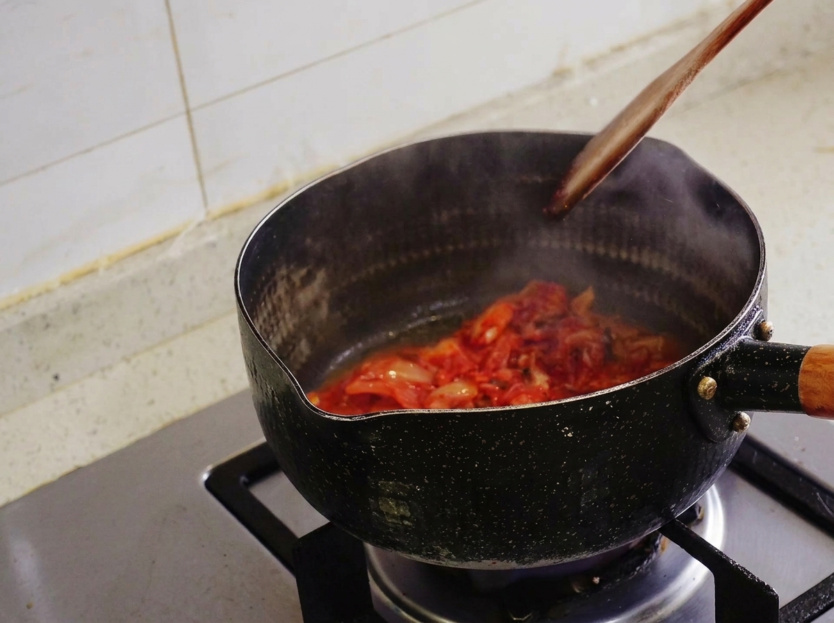 Kimchi podsmażane drewnianą łopatką w czarnym garnku, z którego unosi się para.