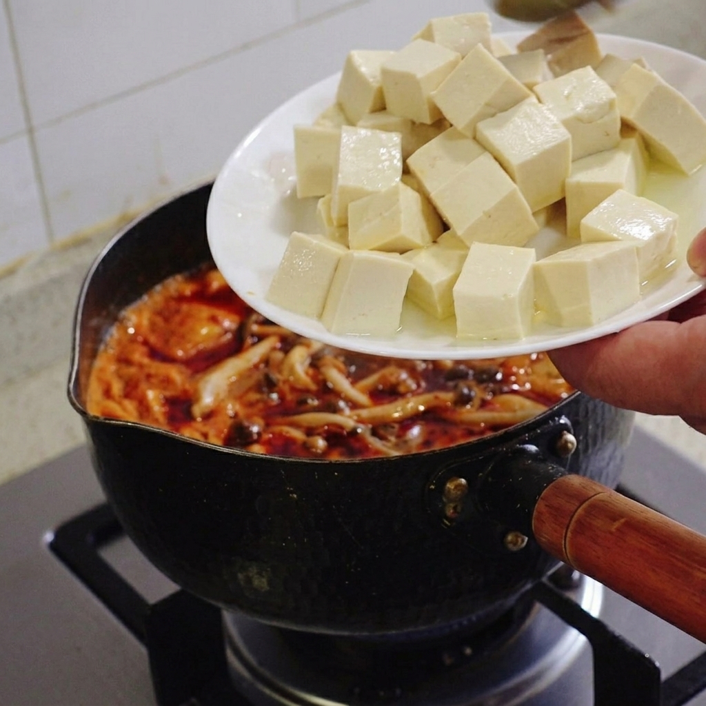 Dłoń trzymająca biały talerz i zsuwająca równe kostki miękkiego białego tofu do mrugającego garnka z zupą kimchi.