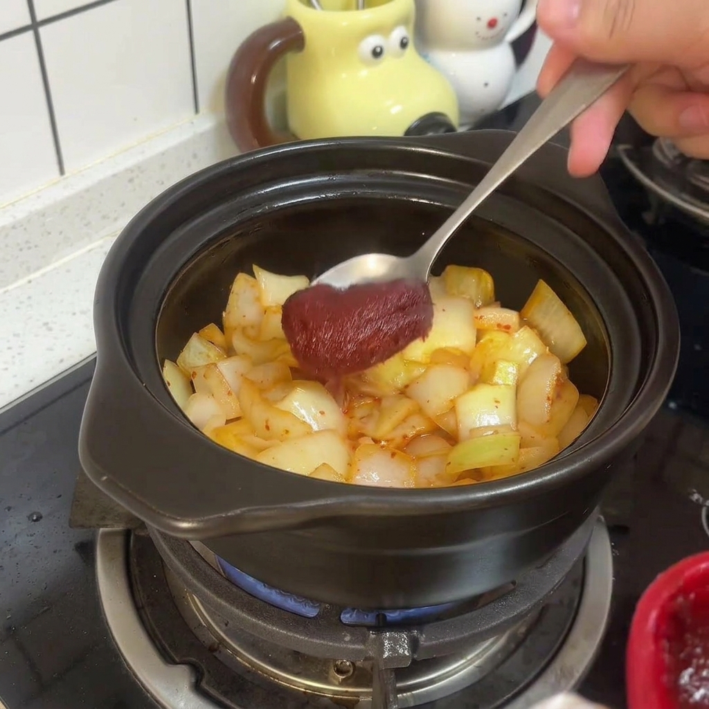 Metalowa łyżka mieszająca gęstą czerwoną koreańską pastę chili z podsmażonym kimchi i cebulą w glinianym naczyniu.