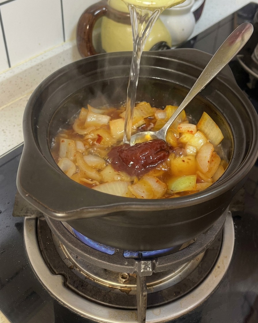 Woda wlewana do glinianego garnka z podsmażoną cebulą, kimchi i czerwoną pastą chili.
