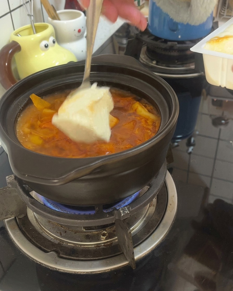 Łyżka nabierająca duże kawałki miękkiego tofu do bulgoczącego czerwonego bulionu w glinianym garnku.