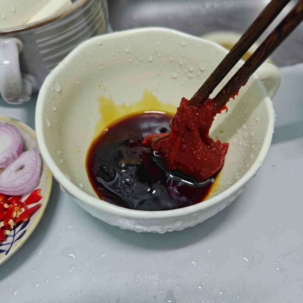 Drewniane pałeczki trzymające dużą porcję gęstej, czerwonej koreańskiej pasty chili nad białą ceramiczną miską z ciemnymi sosami.