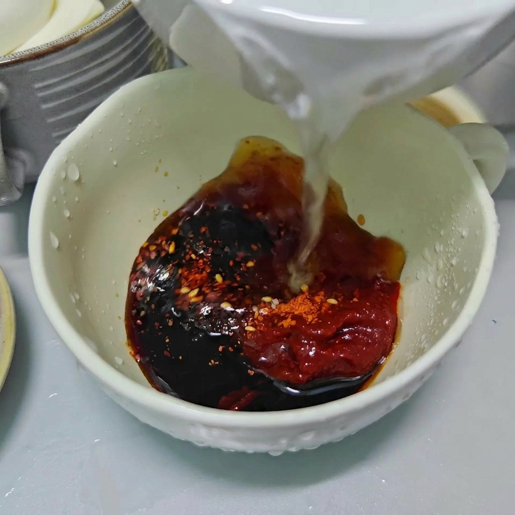 Czysta woda wlewana do białej miski wypełnionej ciemnym sosem sojowym, czerwoną pastą chili i płatkami chili.