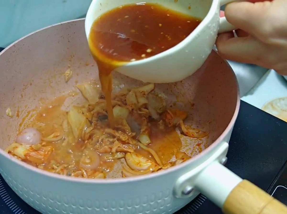 Osoba wlewająca czerwonawy sos z białego kubka na różową patelnię z podsmażonym kimchi i cebulą.