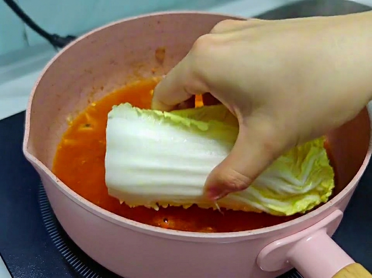 Dłoń ostrożnie układająca duży kawałek świeżej kapusty pekińskiej w różowej patelni z bulgoczącym bulionem kimchi.