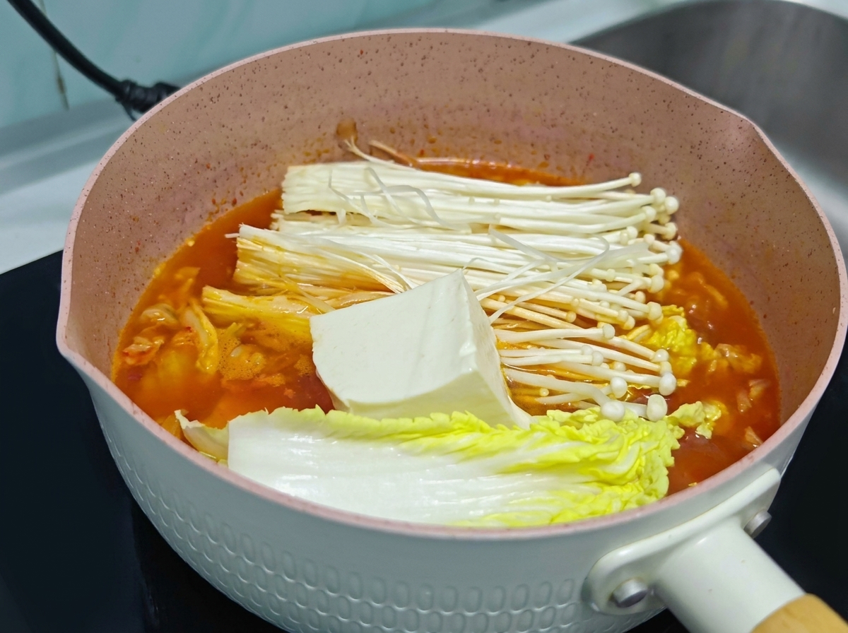 Duży kawałek świeżego białego tofu ułożony na grzybach enoki i kapuście w garnku z pomarańczową zupą kimchi.