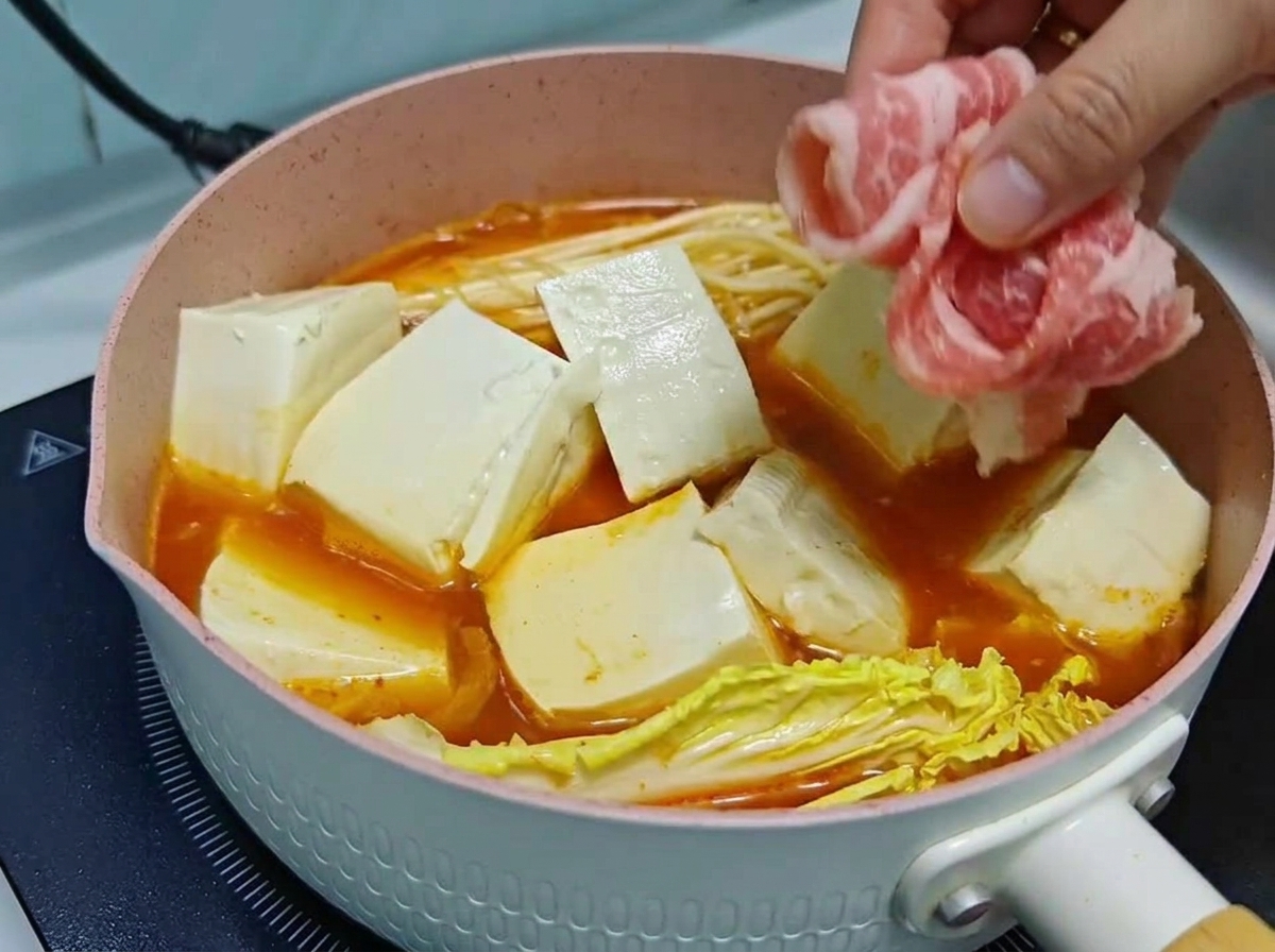 Dłoń ostrożnie układająca cienkie plastry surowej wołowiny na kostkach tofu i warzywach w gotującym się garnku z zupą kimchi.