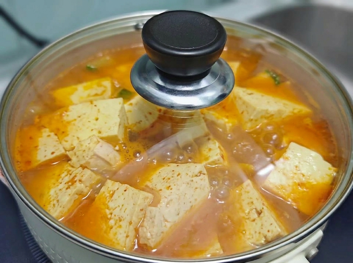 Przezroczysta szklana pokrywka przykrywająca garnek z bulgoczącą koreańską zupą kimchi z wołowiną i tofu na kuchence.