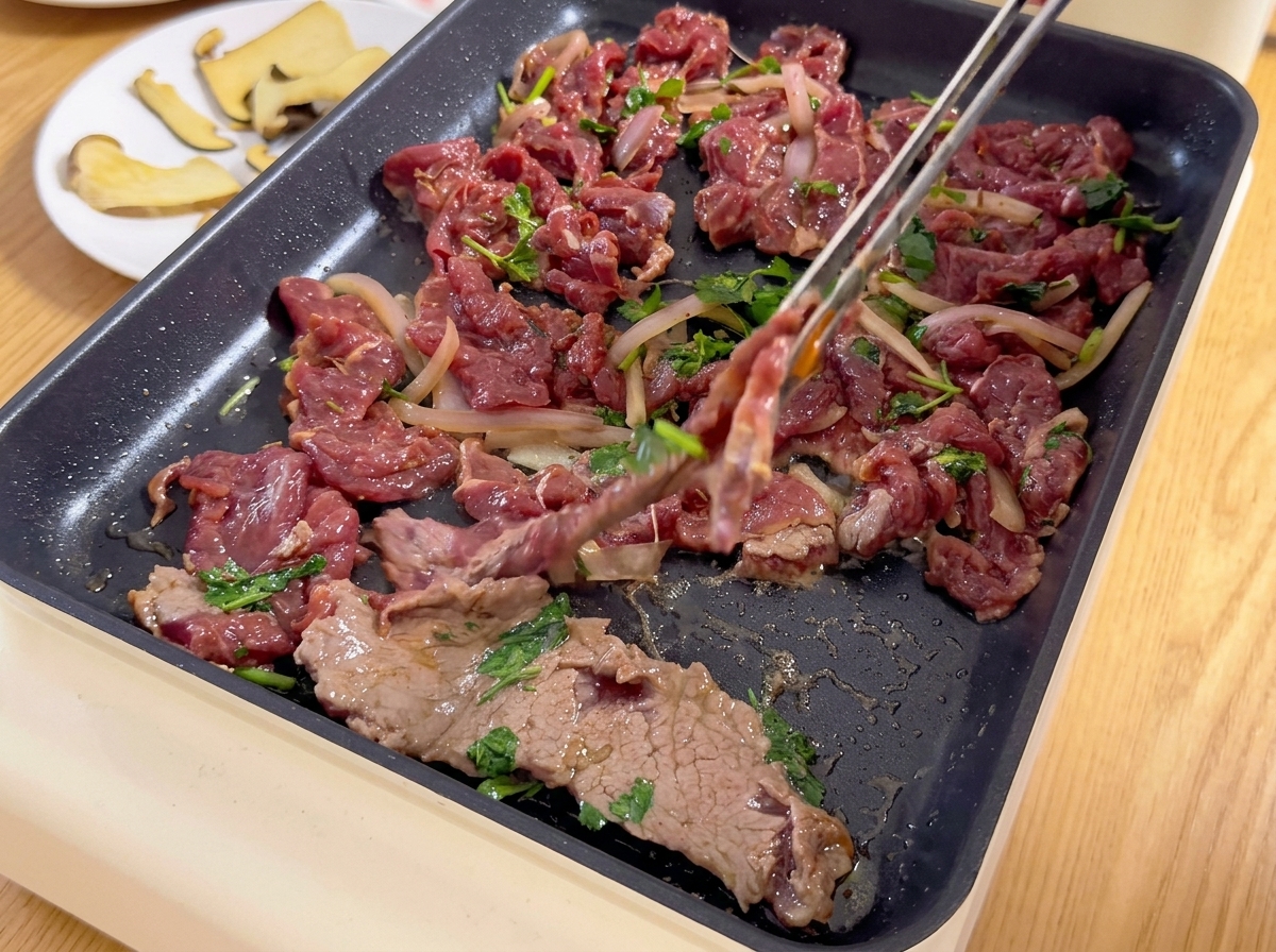 Cienko pokrojona, zamarynowana wołowina z cebulą i kolendrą nakładana szczypcami na gorący grill elektryczny.