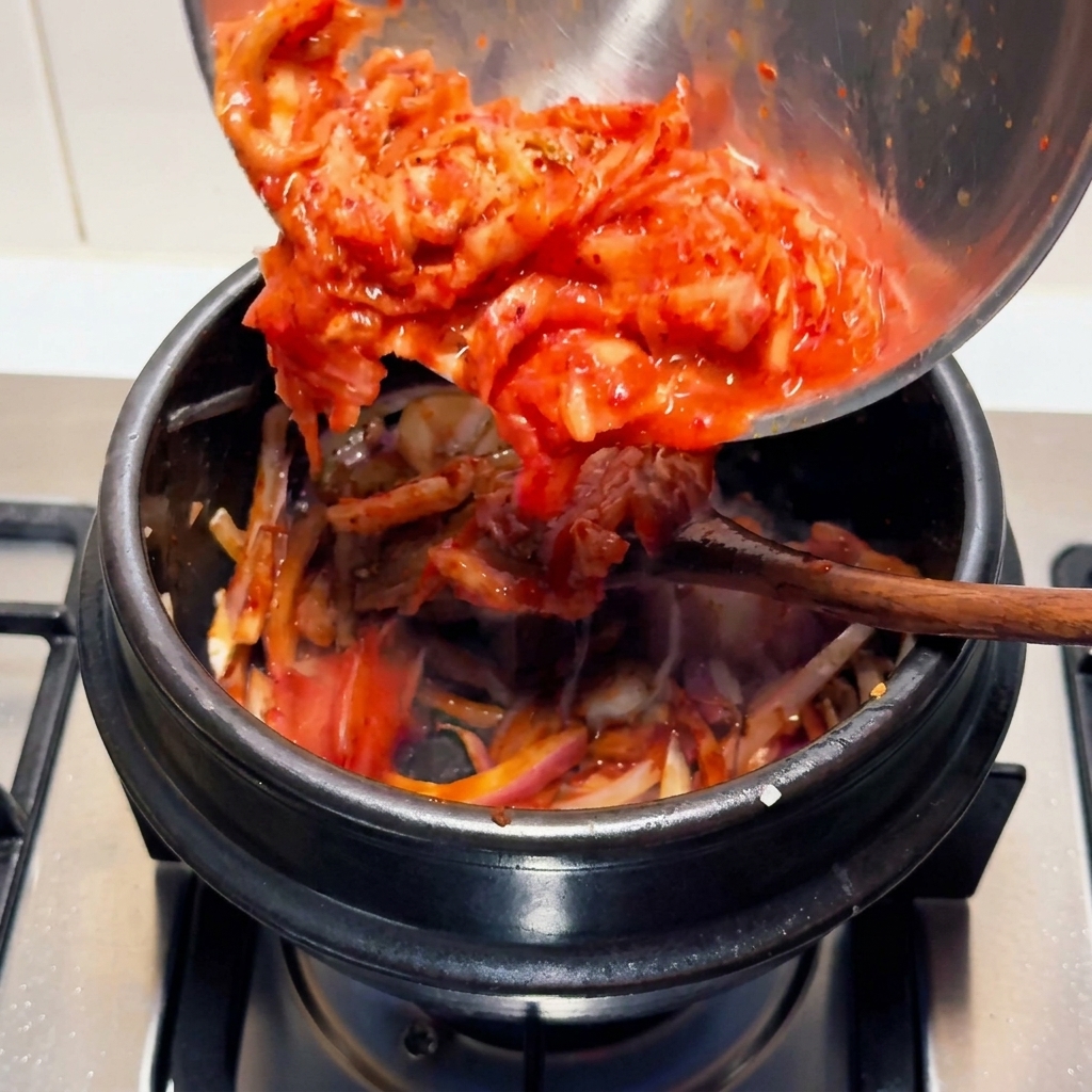 Jasnoczerwone, posiekane kwaśne kimchi przekładane z metalowej miski do czarnego kamiennego garnka.