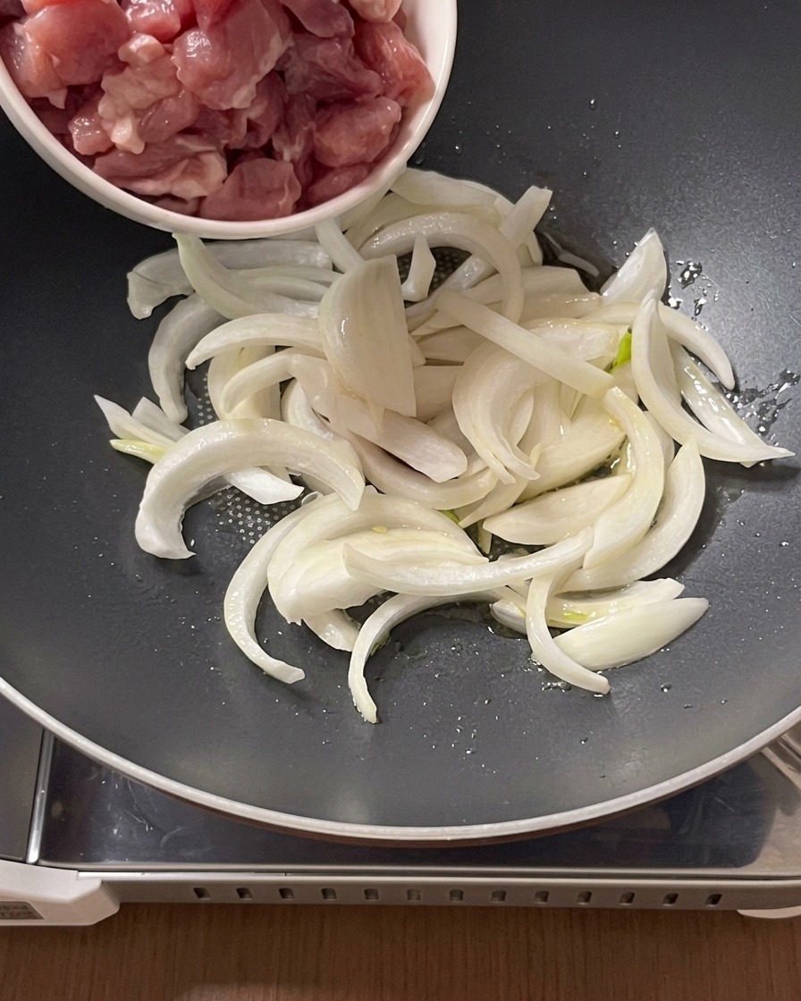 Surowe kawałki wieprzowiny przekładane z małej miski na patelnię z pokrojoną cebulą i olejem.