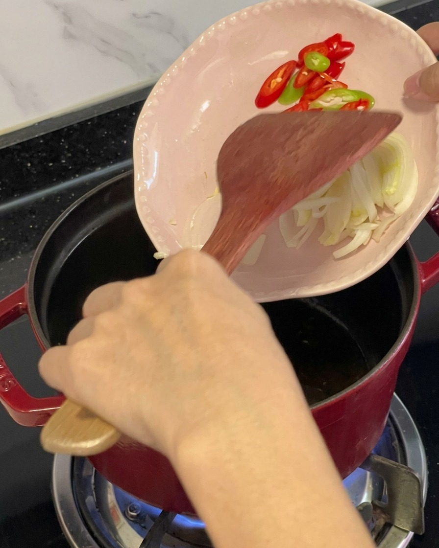 Pokrojone czerwone i zielone chili oraz składniki aromatyczne zsuwane z małego różowego talerzyka do naoliwionego czerwonego garnka za pomocą drewnianej łopatki.