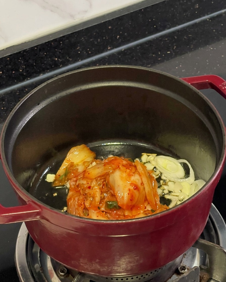 Posiekane czerwone kimchi, pokrojony czosnek i dymka smażące się na oleju w garnku.