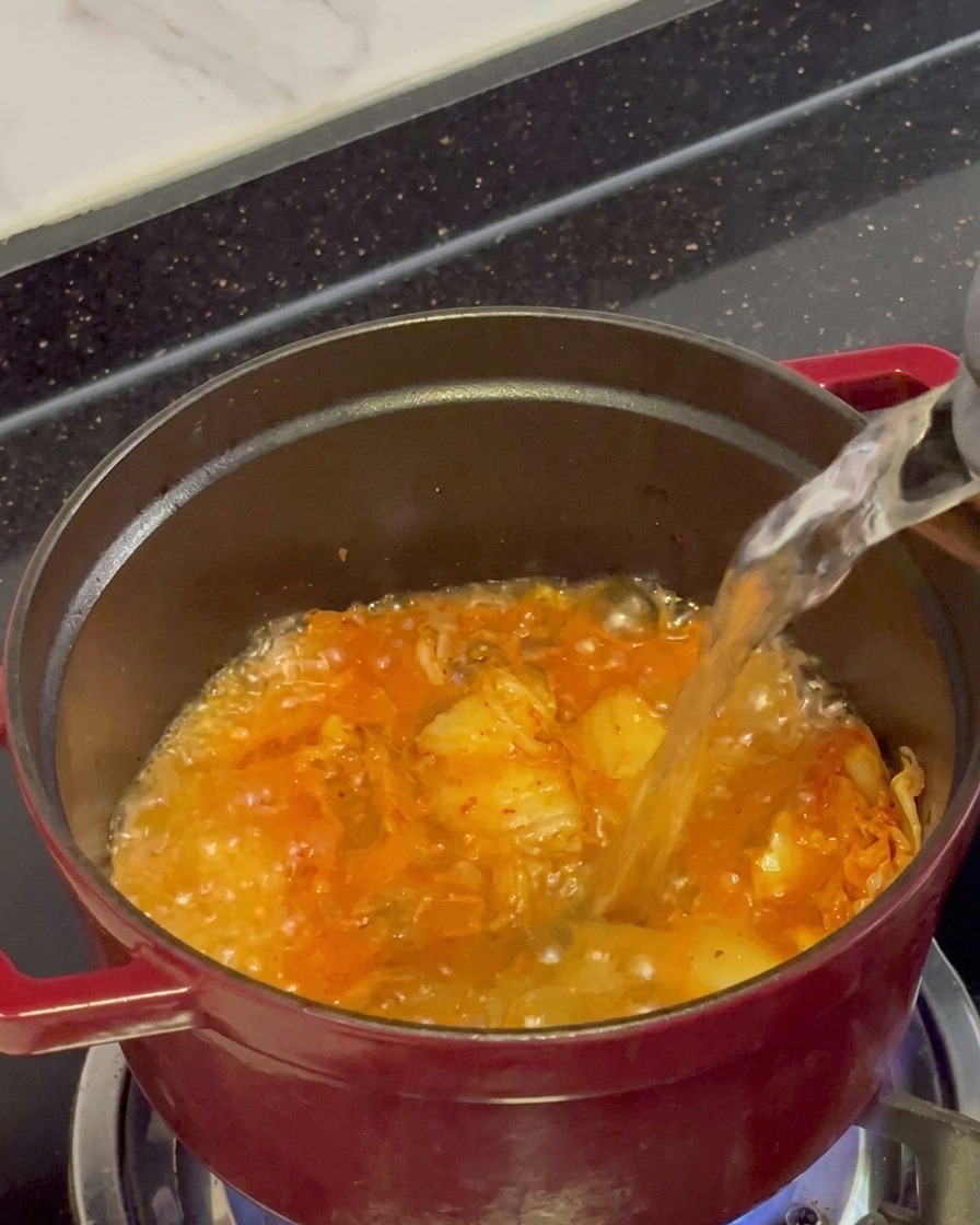 Czysty wrzątek wlewany z czajnika do czerwonego garnka wypełnionego bulgoczącym, podsmażonym kimchi.