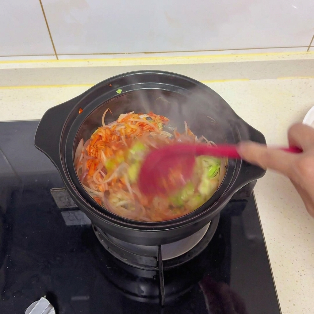Mieszanie warzyw i kimchi w czarnym garnku czerwoną szpatułką w celu rozprowadzenia soli i sosu sojowego.