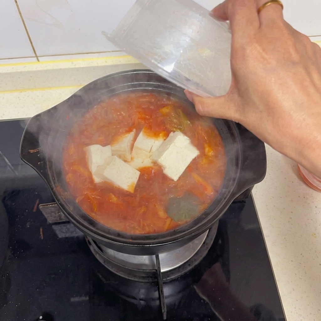Dłoń delikatnie przekładająca duże kostki białego tofu z przezroczystego pojemnika do gotującej się zupy z kimchi.