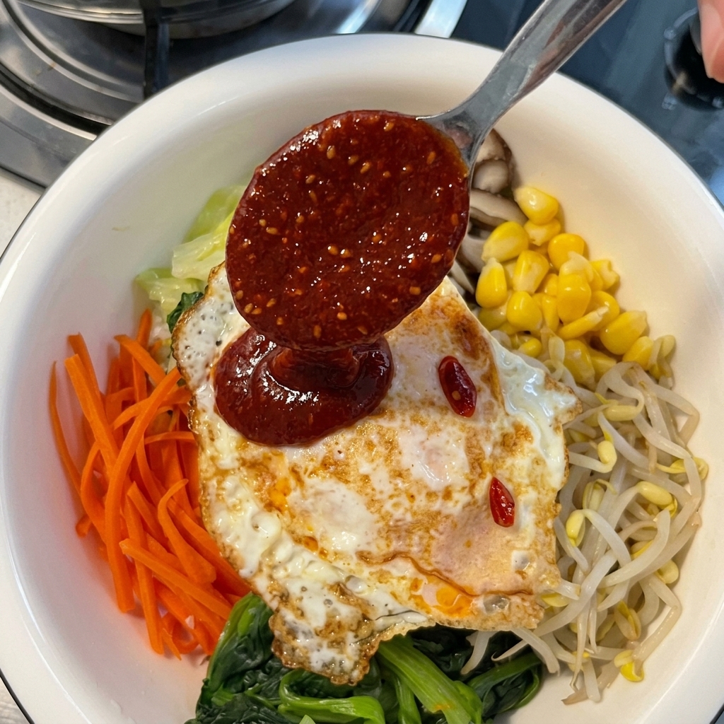 Polewanie gęstym, czerwonym sosem chili z metalowej łyżki jajka sadzonego ułożonego na kolorowych warzywach do bibimbap.