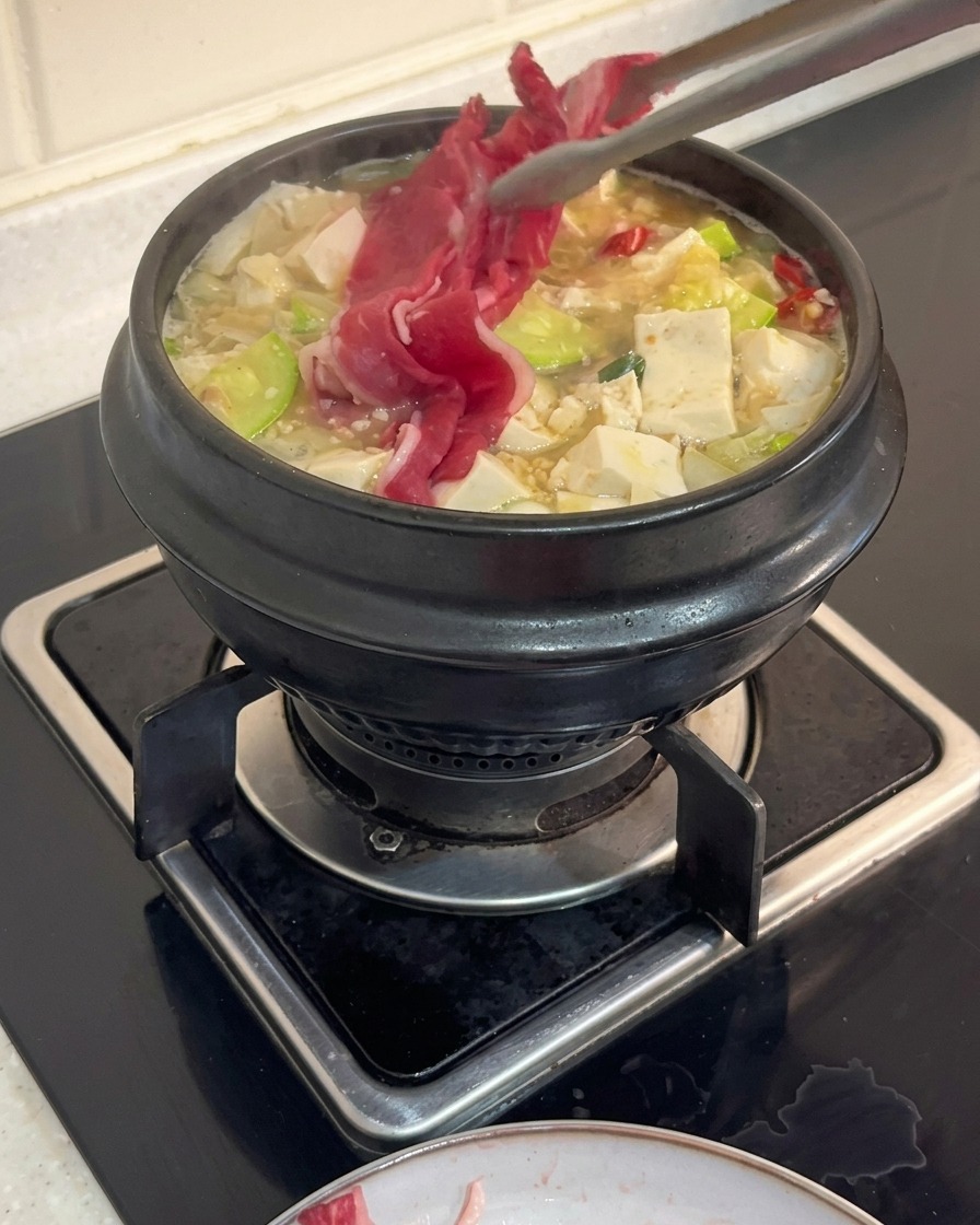 Szczypce kuchenne nakładające cienkie plastry surowego mostka wołowego do wrzącego koreańskiego gulaszu z tofu i cukinią.