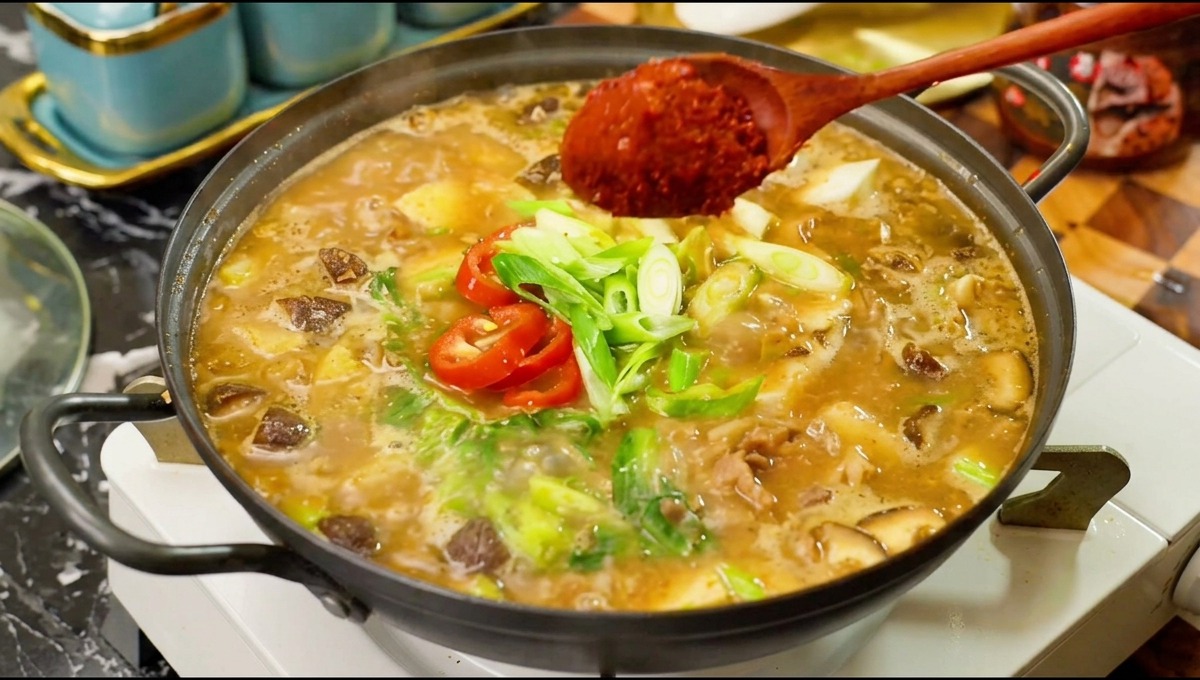 Drewniana łyżka dodająca dużą porcję jasnoczerwonej papryki chili do bulgoczącego garnka z zupą sojową, wołowiną, grzybami i świeżym chili.