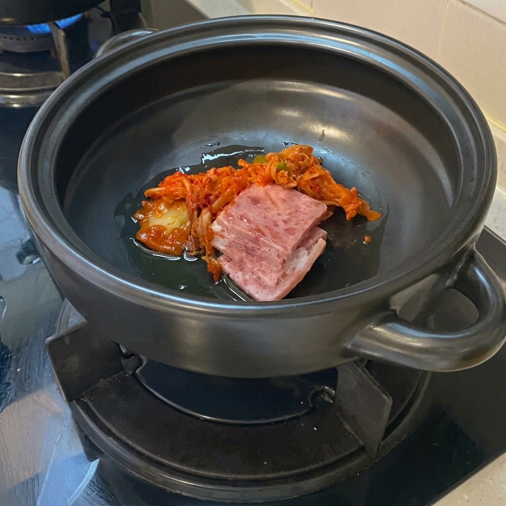 Plastry surowego boczku i kawałki wyrazistego, posiekanego kimchi na oleju na dnie ciemnego glinianego garnka ceramicznego.