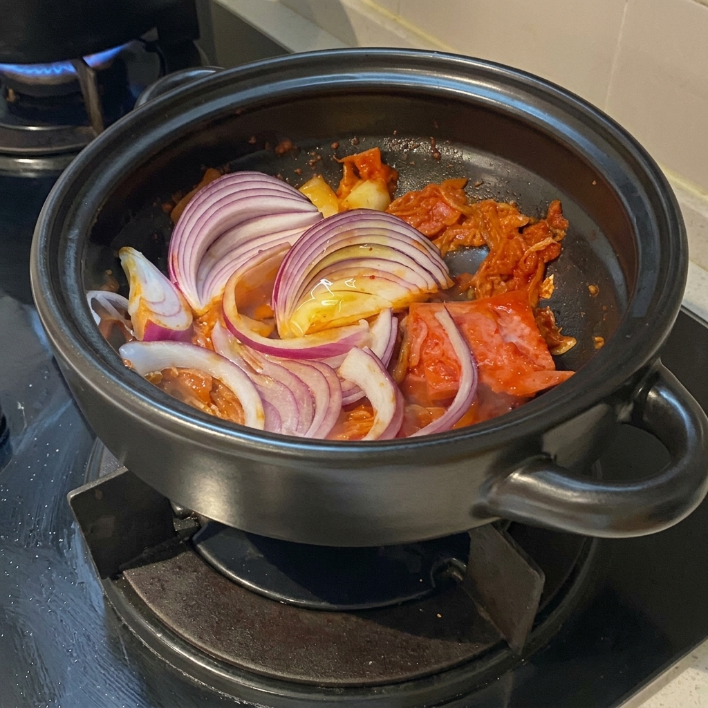 Grube piórka intensywnej czerwonej cebuli ułożone na podsmażonym kimchi z boczkiem wewnątrz glinianego garnka.