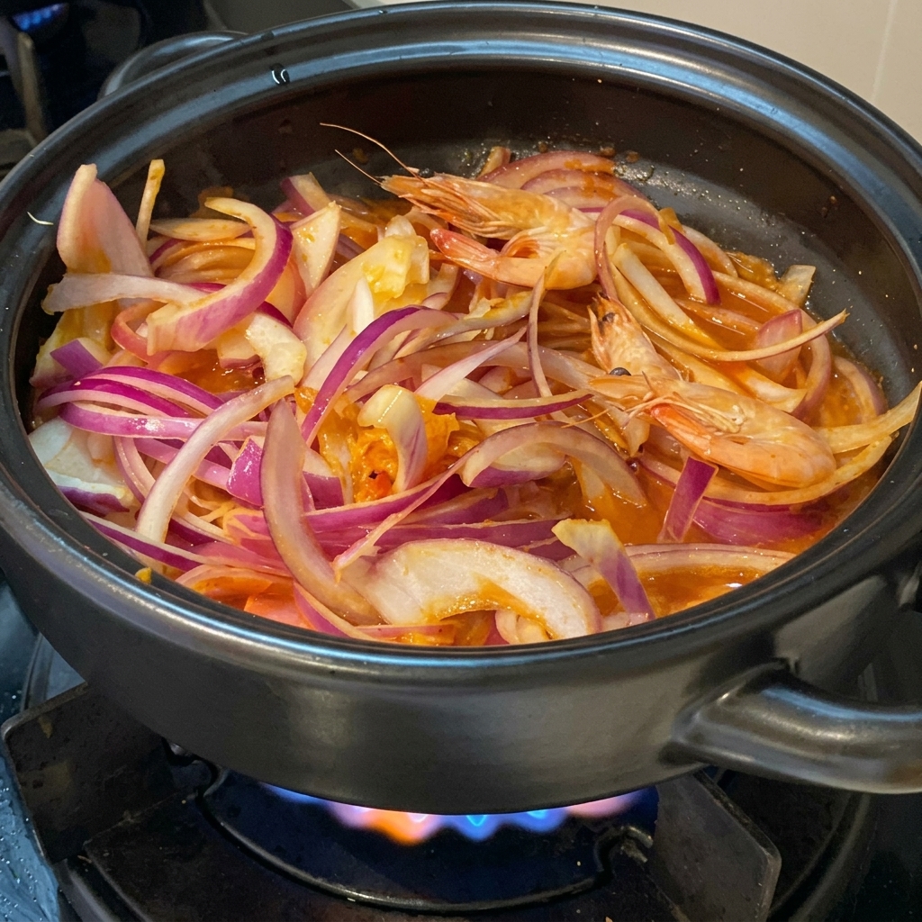 Świeże, całe różowe krewetki ułożone pomiędzy piórkami czerwonej cebuli i kimchi w czarnym glinianym garnku na kuchence.