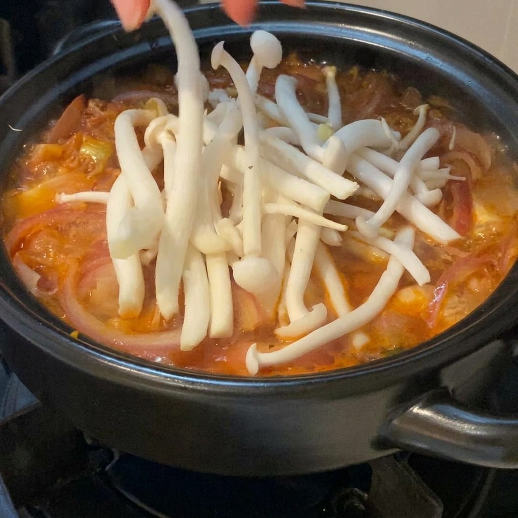 Świeże, białe grzyby shimeji delikatnie układane dłonią na powierzchni bulgoczącej, czerwonej zupy kimchi w czarnym garnku.