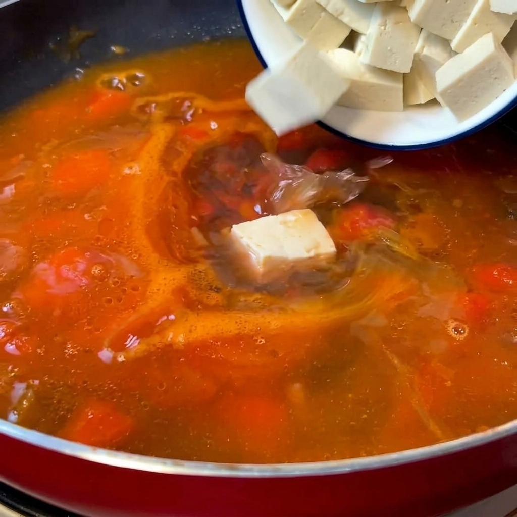 Kostki białego tofu wrzucane do patelni z gotującą się pomidorową bazą zupy.