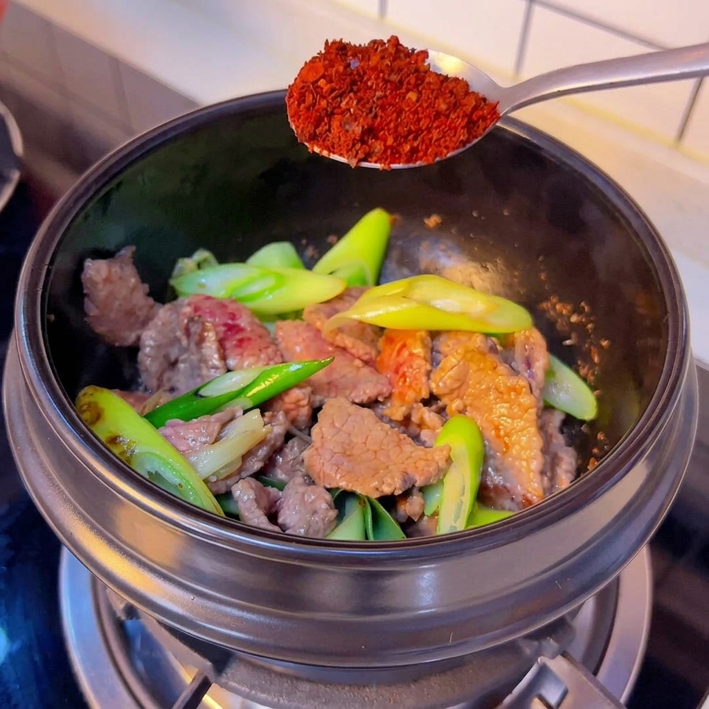 Srebrna łyżka z grubo mielonym czerwonym chili nad czarnym garnkiem ze smażoną wołowiną i dymką.